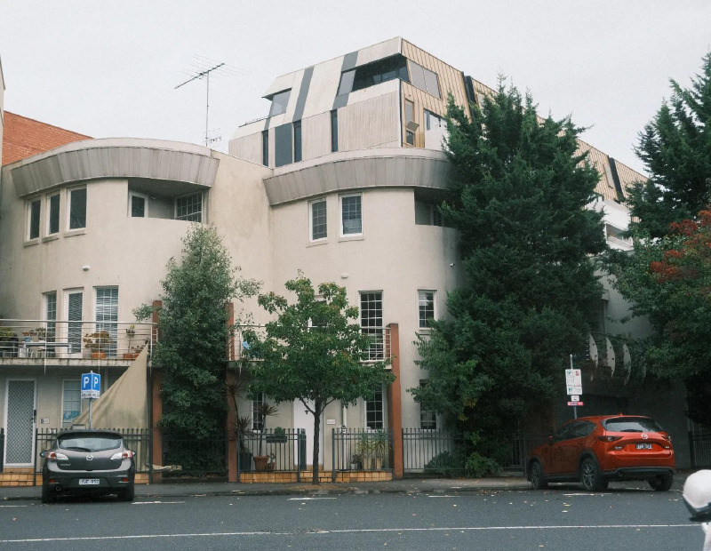 A photograph of a two-storey apartment building, the front is rounded with a square structure on the top level. A number of trees grow next to it, the tallest is the height of the building. The dominant colors are cream, green and red.