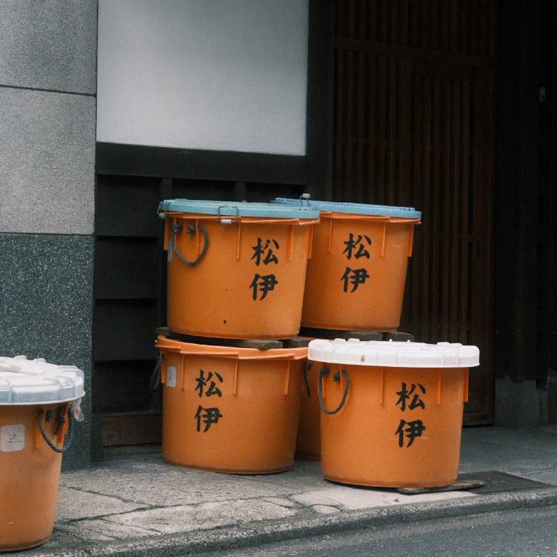 A photograph of a stack of round plastic tubs sitting next to a building. The tubs are bright orange, with white and blue lids. The name 松伊 is written on the side in kanji.