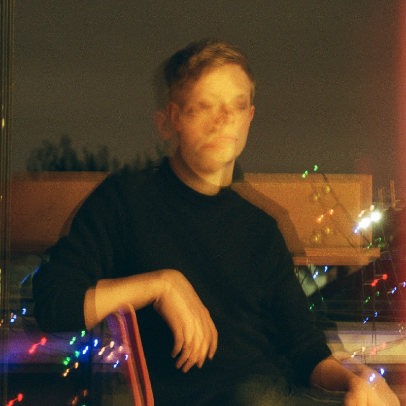 A self-portrait of River sitting on a chair on a balcony at night. It is blurry, as if two very similar images were laid over each other.