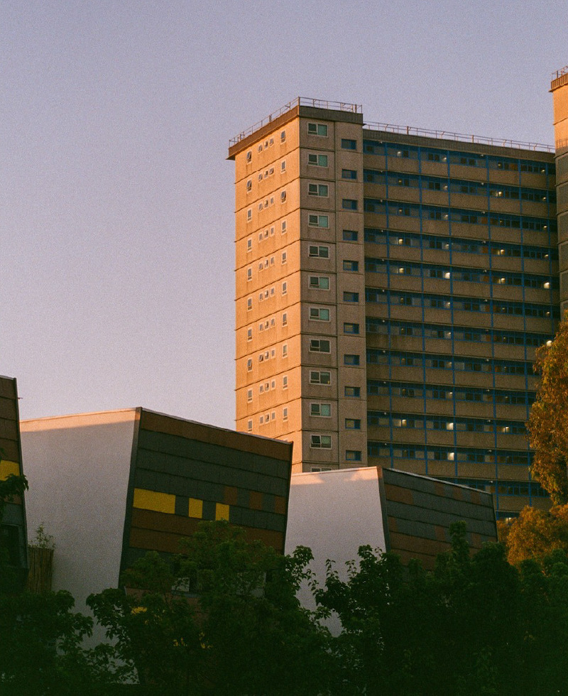 A photograph of a tall apartment block, taken at sunset. It is framed by a pink-blue, cloudless sky. One side is lit up orange from the sunset.